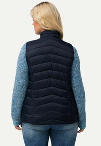 Marineblaue Steppweste mit Zickzacksteppung, hohem Kragen, kombiniert mit einem blauen strukturierten Strickpullover. Die Rückansicht zeigt eine taillierte Silhouette.