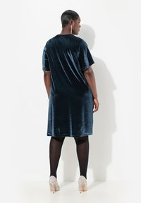 Gaun beludru dalam warna teal tua, dengan potongan longgar, lengan pendek, dan hem setinggi lutut. Dipasangkan dengan sepatu hak tinggi yang berkilau dan dihiasi.