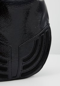 Gros plan d'un coin de sac à main en cuir verni noir avec des détails de couture incurvée sur un fond clair.