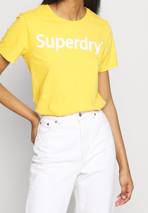 Kobieta w żółtej koszulce Superdry włożonej do białych spodni z wysokim stanem, z jedną ręką opartą na biodrze.