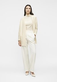 Γυναίκα που στέκεται, ντυμένη με κρεμ ζιβάγκο, ανοιχτή ζακέτα, φαρδιά παντελόνια και μυτερά ψηλοτάκουνα σε λευκό φόντο.