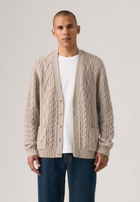 Man wearing beige cable-knit cardigan over white t-shirt and blue jeans, standing against a plain light background.