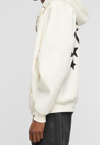 Sudadera con capucha en blanco roto, con estampados de estrellas negras a lo largo de la manga izquierda, puños acanalados y un bolsillo tipo canguro. Tejido suave y texturizado.