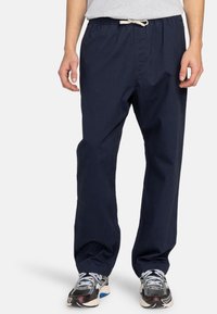 Pantalon bleu marine léger à cordon de serrage avec une coupe décontractée et une texture subtile. Porté avec des chaussures de sport grises, illustrant un style décontracté.