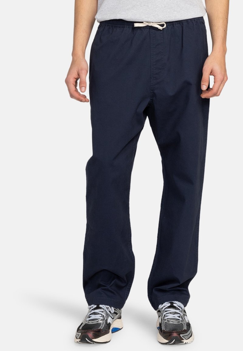 Pantalon bleu marine léger à cordon de serrage avec une coupe décontractée et une texture subtile. Porté avec des chaussures de sport grises, illustrant un style décontracté.