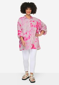 Maglietta leggera tie-dye rosa con maniche a palloncino, chiusura anteriore con bottoni; abbinata a pantaloni bianchi aderenti e sandali chunky con cinturini.