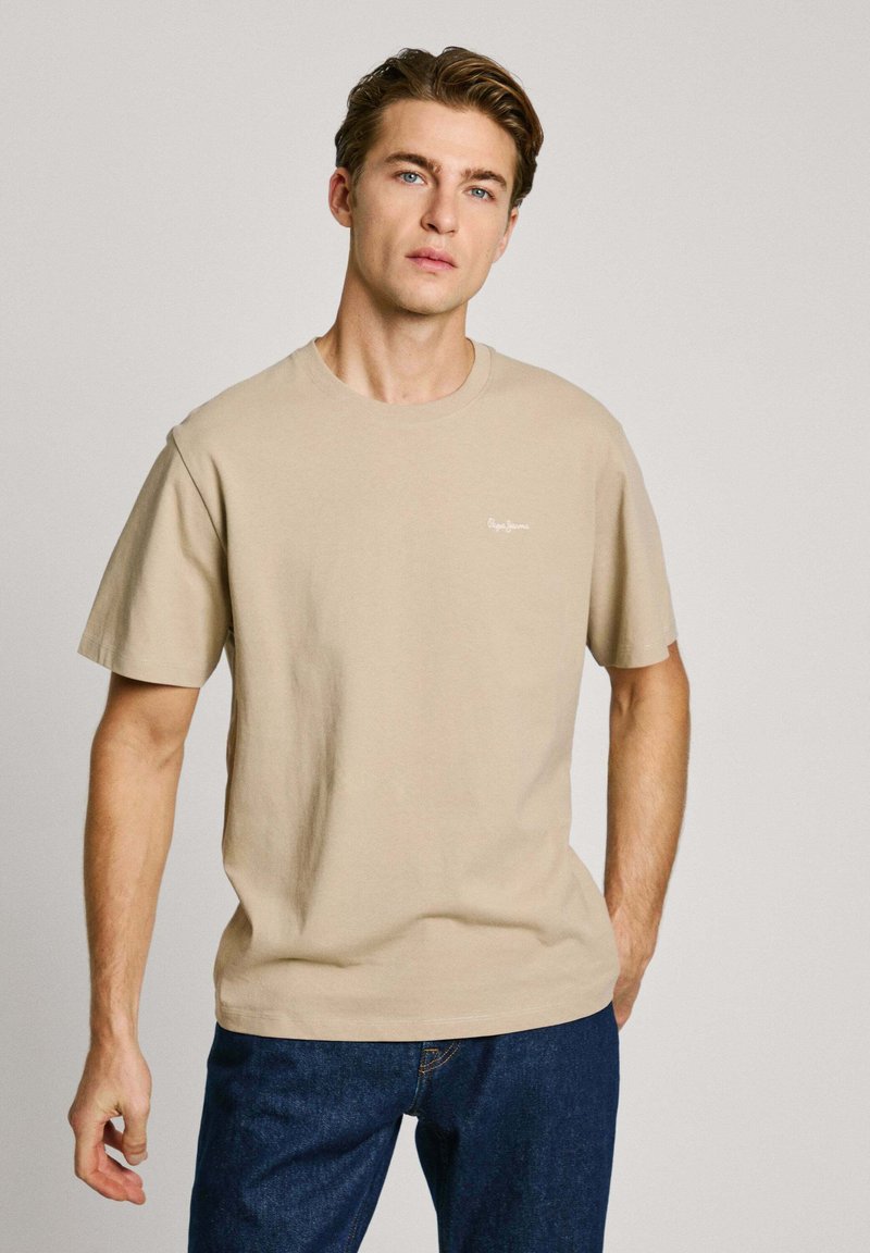 Beige cotton t-shirt with short sleeves, crew neck, and small white logo on the left side. Paired with blue jeans. Simple and classic design.