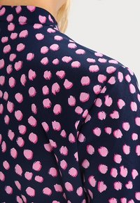 Marineblaue Stoff mit pinken Pinselstrich-Punkten, der eine glatte Textur und ein hochgeschlossenes Detail aufweist, von hinten betrachtet.