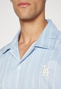 Camisa azul-claro com riscas brancas verticais, com colarinho e um pequeno logótipo bordado no lado esquerdo do peito. Tecido com textura suave.