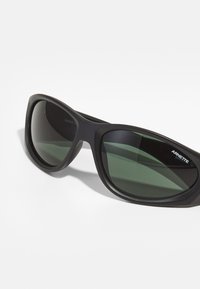 Occhiali da sole neri con finitura opaca, lenti ovali grandi di colore verde scuro e un marchio prominente sul lato. Design resistente con curve sottili.
