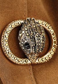 Fibbia in metallo color oro con design dettagliato a forma di artiglio decorato con gemme chiare e nere, su sfondo in camoscio color cuoio.