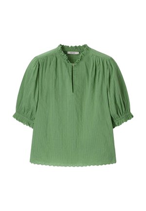 Vrouw met lang bruin haar, die een groene blouse met geschulpte randen en lichtblauwe spijkerbroek draagt, poseert met haar armen boven haar hoofd.