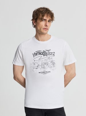 Uomo con capelli ricci che indossa una maglietta bianca con un'illustrazione vintage di una motocicletta e le parole "Vintage Motors" e "Motorcycles".