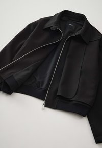 Zwarte bomberjack met een ritsluiting, gestructureerde buitenkant en gladde voering. Heeft een klassieke kraag en ribgebreide manchetten.