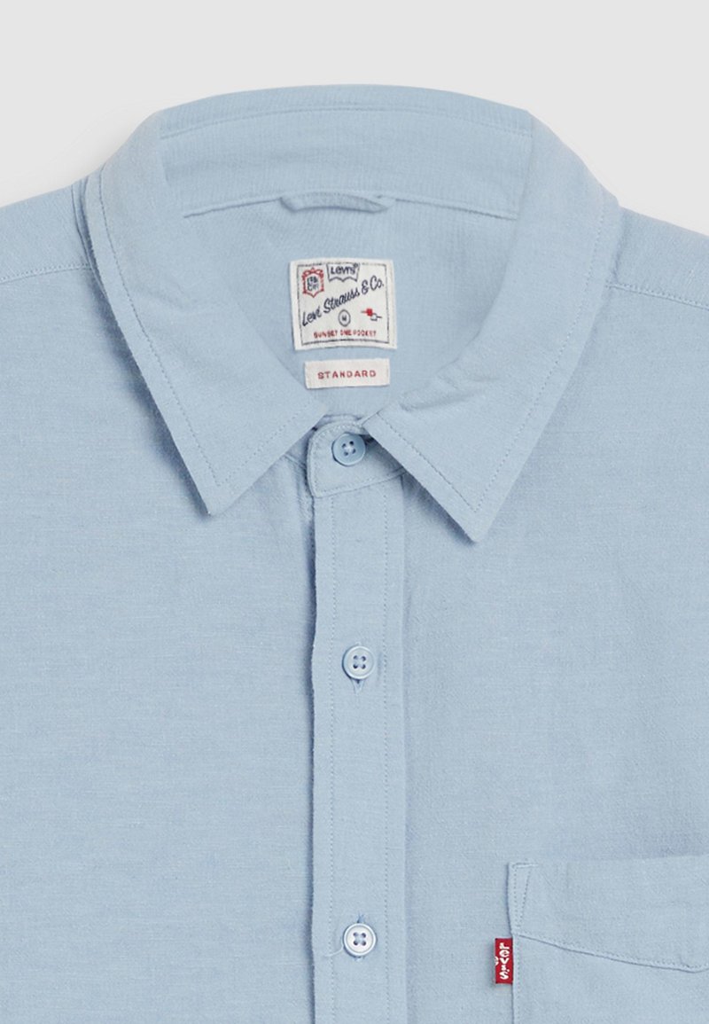 Lichtblauwe button-up shirt met kraag, voorzien van twee knoopsluitingen, een borstzak en een klein rood logo label. Gladde stof.