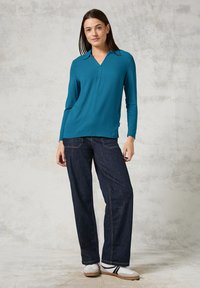 Türkisfarbener Langarm-Oberteil mit V-Ausschnitt und seitlichen Schlitzen, kombiniert mit dunkelblauen weiten Jeans und weißen Sneakers mit schwarzen Akzenten.