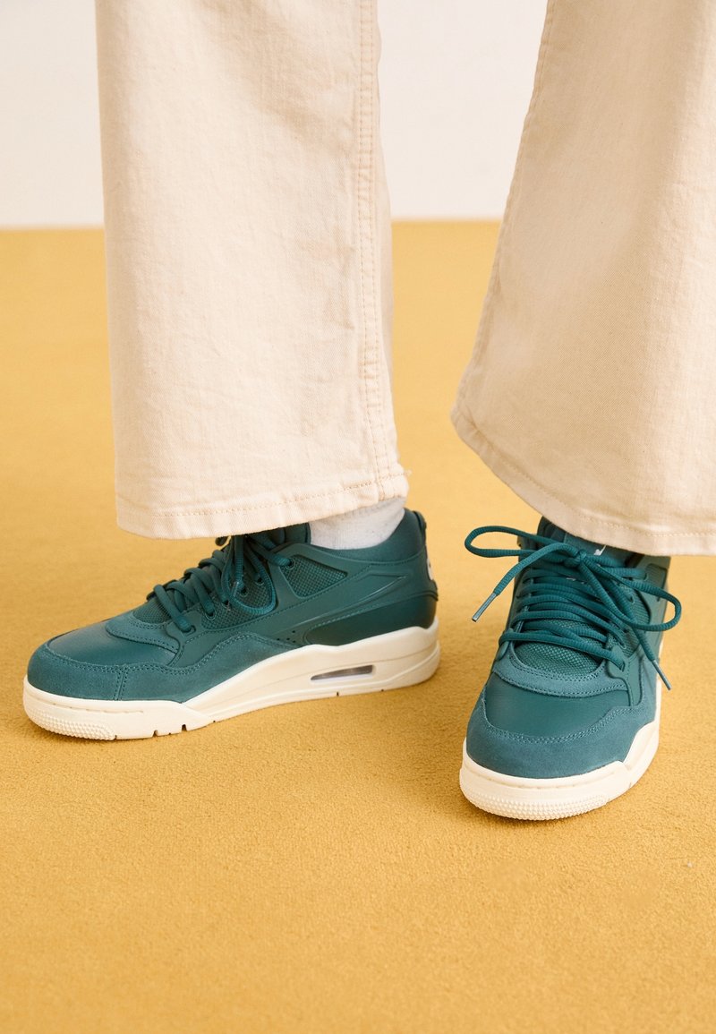 Baskets bleu sarcelle à semelles blanches portées avec des chaussettes blanches et un pantalon beige à jambes larges, debout sur un sol jaune moutarde.