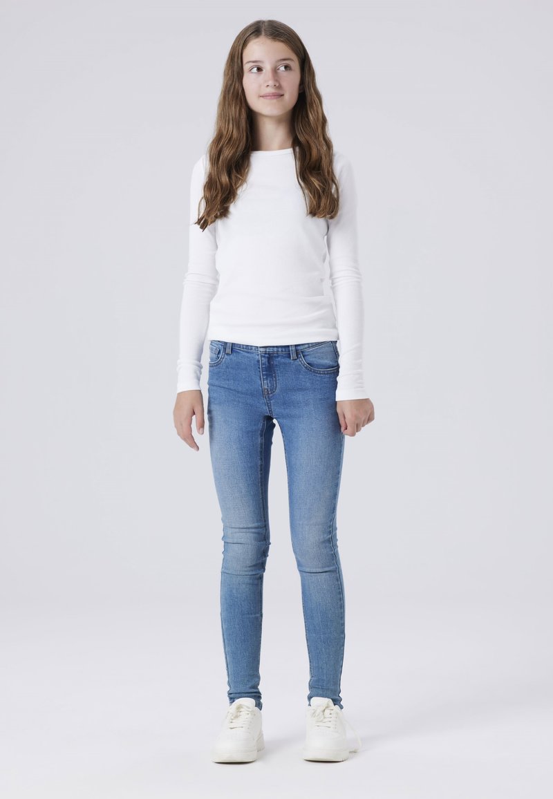 Top blanco de manga larga, jeans ajustados azul claro y zapatillas blancas. El conjunto presenta un diseño sencillo con una textura suave.