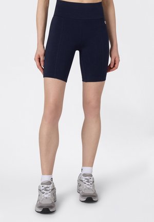 Persona vistiendo pantalones cortos biker azul marino de talle alto, calcetines blancos hasta el tobillo y zapatillas deportivas grises, de pie sobre un fondo blanco.