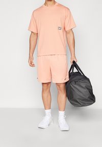 Light pink athletic t-shirt with a small logo, matching shorts, and white sneakers. The individual holds a dark gray duffel bag.
