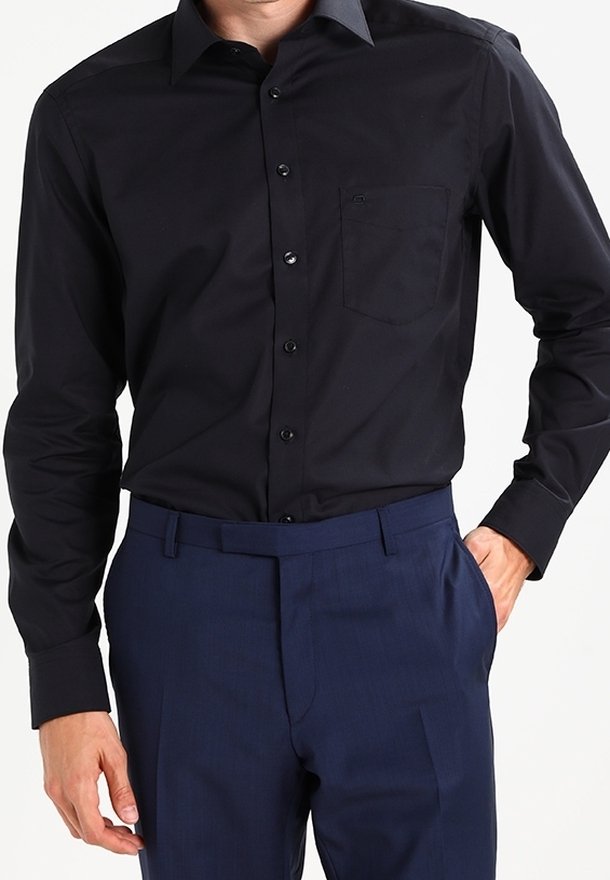 Homme portant une chemise noire boutonnée avec une poche poitrine et un pantalon bleu foncé ajusté, une main dans la poche.