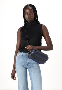 Femeie cu breton lung, purtând un top negru fără mâneci și blugi albaștri deschiși, ținând o geantă crossbody bleumarin și neagră.