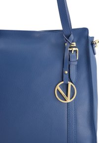 Blauwe leren tote bag met een gestructureerde afwerking, gouden hardware en een kenmerkende ronde logo hanger. Heeft een eenvoudig ontwerp en een platte vorm.
