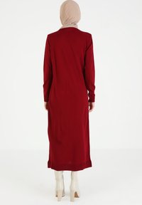 Largo vestido rojo con mangas largas, textura acanalada y corte hasta los tobillos. Llevado con botas de tacón de color claro. Diseño simple, sin patrones visibles.