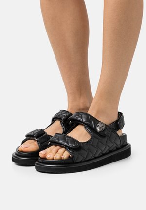 Czarne plecione sandały z dwiema regulowanymi klamerkami, płaską podeszwą i dekoracyjnym srebrnym akcentem na zewnętrznej pasku.