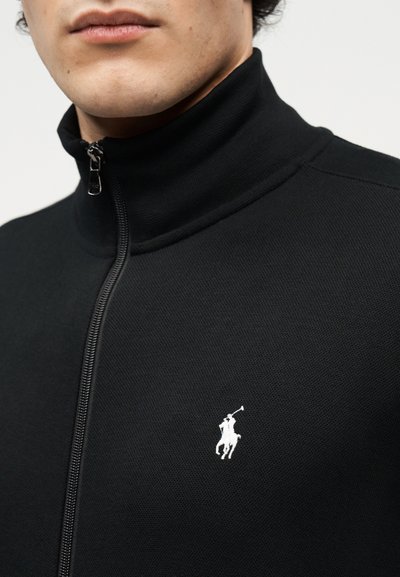 Detailní záběr osoby nosící černou bundu na zip s bílým vyšívaným logem jezdce na pólu na hrudi.