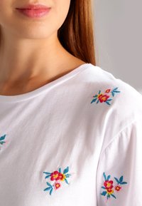 Camiseta de algodón blanca con bordado floral colorido en los hombros, con acentos en rosa, amarillo y azul. Diseño clásico de cuello redondo.
