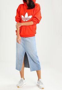 Red hooded sweatshirt with white logo, paired with a knee-length light blue denim skirt featuring a front slit and frayed hem, worn with gray sneakers.