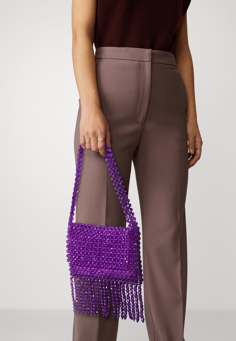 Personne tenant un sac à main violet orné de perles avec des franges, portant un pantalon mauve et un haut sans manches foncé.