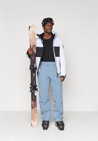 Síelős öltözet, amely magában foglal egy fekete-fehér dzsekit, fekete alapréteget, világoskék nadrágot és fekete sícipőt. Síléceket tart, rajtuk a kötések rögzítve.