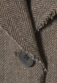 Bruin herringbone-patroon stof met rafelige randen, voorzien van een zwarte knop en subtiele blauwe accenten. Close-up van de textuur van de stof.