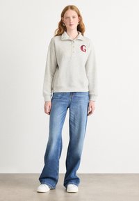 Grå sweatshirt med krage, tre knappar och en röd bokstav "G"-logotyp. Kombinerad med ljusblå utsvängda jeans och vita sneakers.