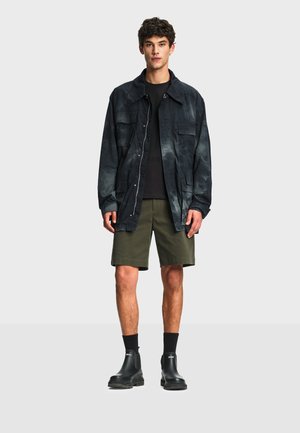 Jeune homme portant une veste surdimensionnée foncée, une chemise noire, un short vert olive, des chaussettes noires et des bottines noires à enfiler, debout devant un fond uni.