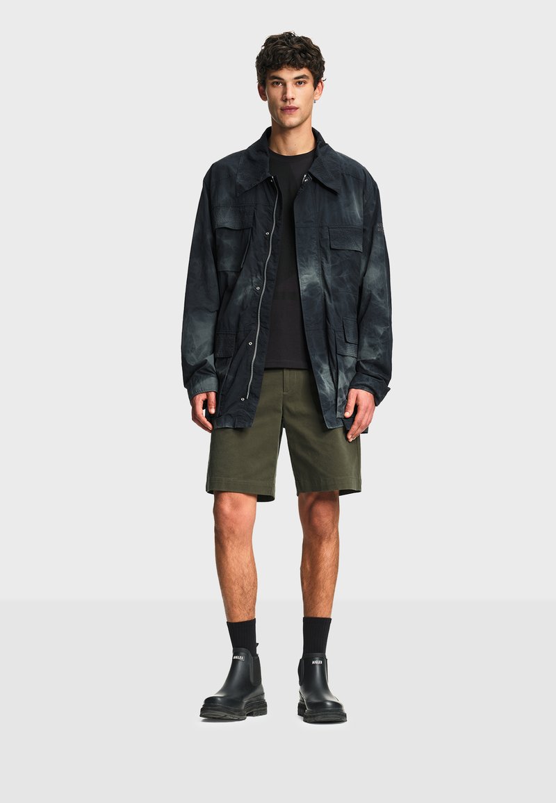 Jeune homme portant une veste surdimensionnée foncée, une chemise noire, un short vert olive, des chaussettes noires et des bottines noires à enfiler, debout devant un fond uni.
