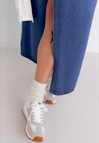 Falda de mezclilla con una abertura lateral, combinada con zapatillas deportivas blancas con detalles grises y suelas de goma marrón, y calcetines crema acanalados.