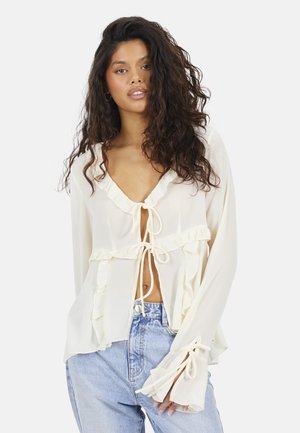 Donna con capelli lunghi e ricci, che indossa una camicetta color crema con balze legata sul davanti e jeans larghi azzurri, su sfondo bianco.