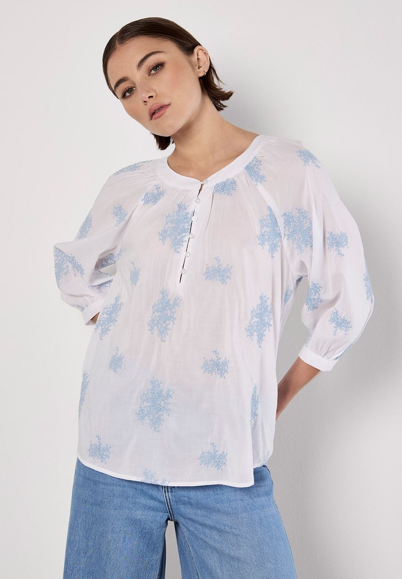 Vit blus med ljusblå blommönsterbroderi, rund hals, knapppdetailj och trekvartsärmar. Mjuk tyg med en avslappnad passform.