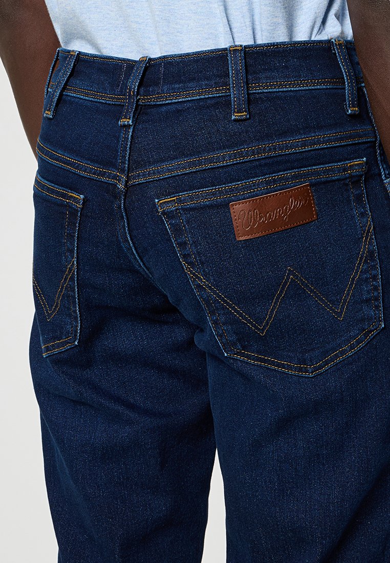 Granatowe dżinsy z charakterystycznym podwójnym ściegiem na tylnych kieszeniach oraz brązową skórzaną metką z wytłoczoną nazwą "Wrangler".