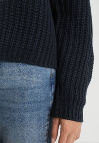 Ciemnogranatowy sweter wykonany na drutach z fakturowanym wzorem prążkowanym i szerokimi mankietami, w zestawieniu z jasnoniebieskimi dżinsami.