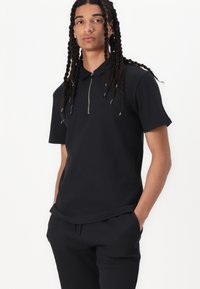 Svart polo skjorte med korte ermer, med glidelås foran og ribbestruktur. Brukt med matchende svarte bukser, minimalistisk design.