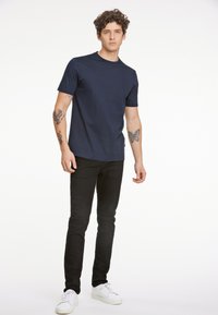 Marineblauw T-shirt met korte mouwen, ronde hals, aansluitende pasvorm, gecombineerd met smalle zwarte jeans en witte sneakers. Toont zichtbare tatoeages op de armen.