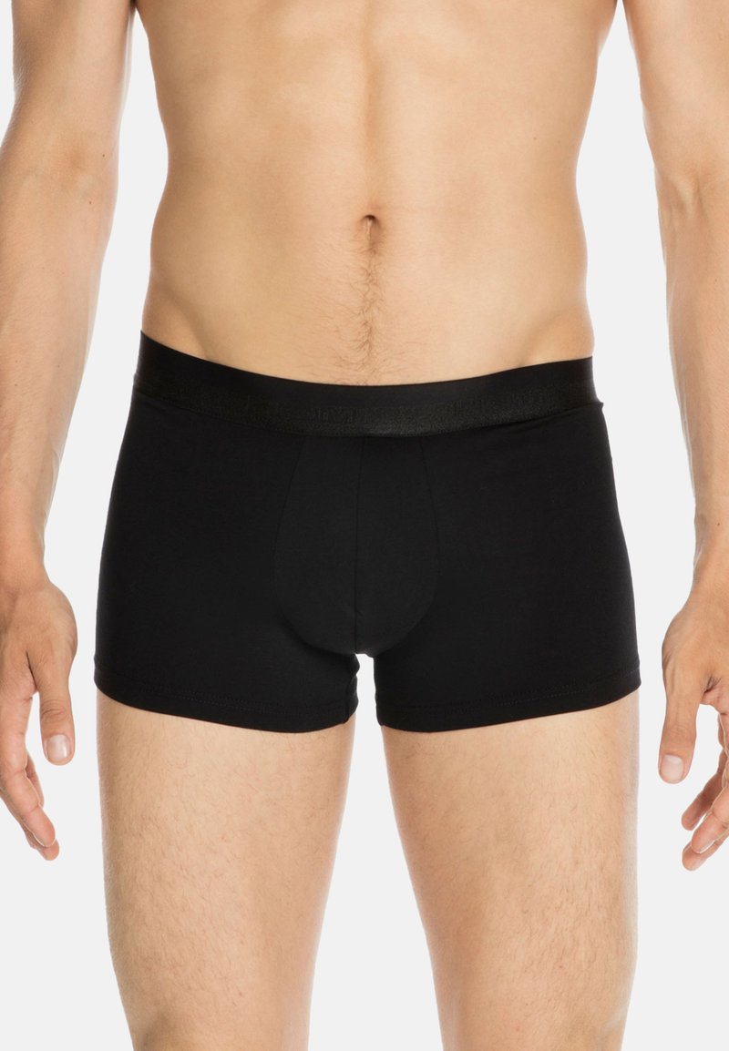 Mies torso, joka on pukeutunut yksinkertaisiin mustiin boxereihin, seisoo kädet rentoina sivuilla valkoista taustaa vasten.