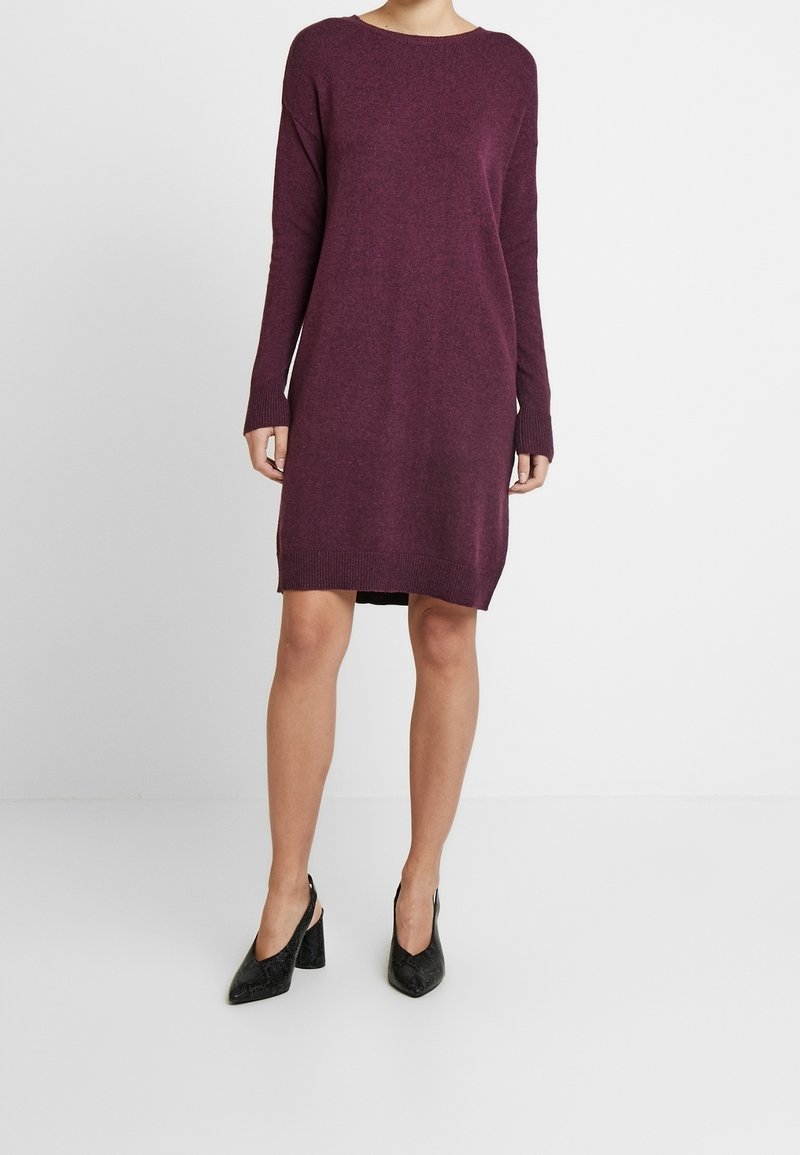 Femme portant une robe tricotée violette à manches longues et longueur genoux avec des chaussures à talons hauts noires à bride arrière, debout devant un fond uni.