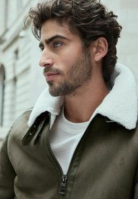 Chaqueta de ante verde oliva con cuello de piel sintética blanca, cierre de cremallera en el frente y detalles de botones a presión, llevada sobre una camiseta blanca texturizada.