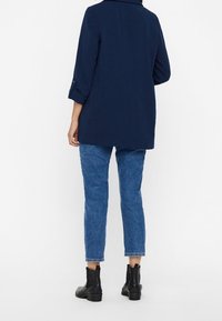 Granatowy, długi marynarka z długim rękawem o dopasowanym kroju, z podwiniętymi mankietami, noszona na jasnoniebieskich dżinsach, uzupełniona czarnymi botkami za kostkę.