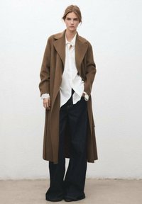 Długi brązowy płaszcz z grubego materiału, zestawiony z białą koszulą i szerokimi ciemnymi spodniami dżinsowymi o szerokich nogawkach. Płaszcz ma wycięty kołnierz.